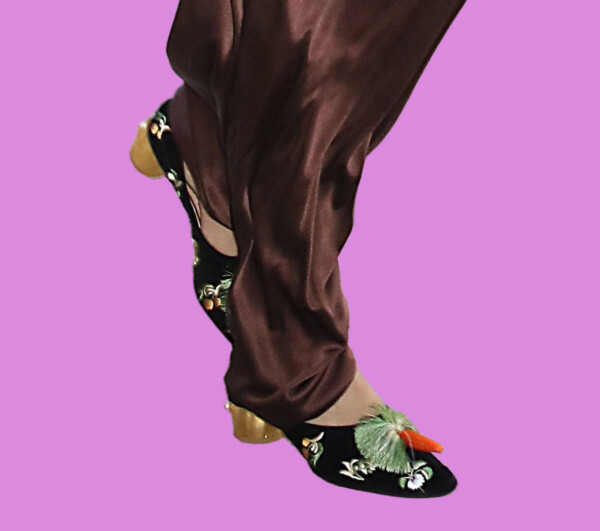 A close-up of a person's foot wearing a black velvet shoe with floral embroidery, paired with brown satin pants, bla bla.