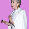 A woman with short blonde hair wearing a white suit and gold earrings, holding a black item, in an urban setting at night, bla bla.