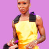 Elegant woman with a shaved head wearing a yellow dress with black shoulder accents, jewelry, and tattoos, standing confidently at an event. bla bla.