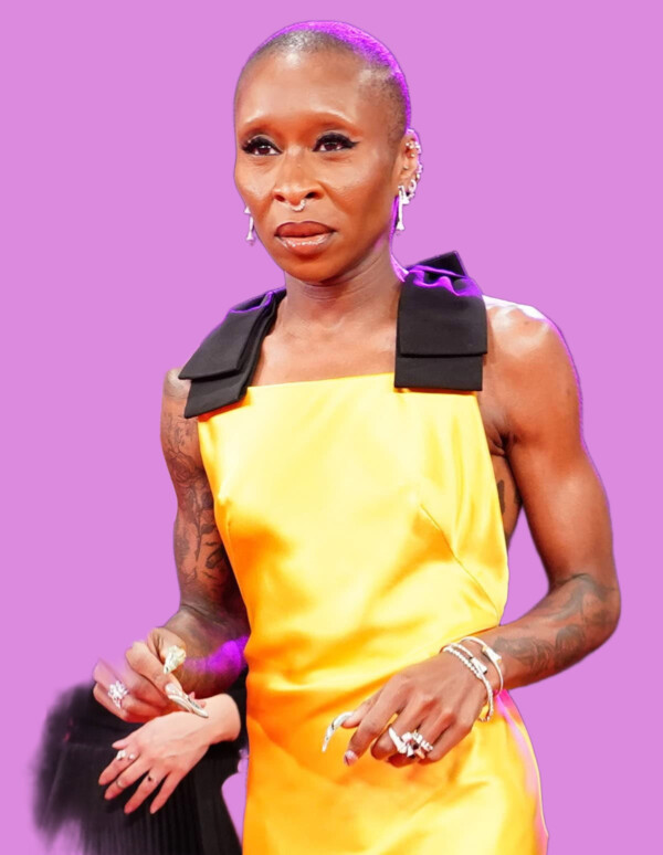 Elegant woman with a shaved head wearing a yellow dress with black shoulder accents, jewelry, and tattoos, standing confidently at an event. bla bla.