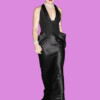 Bla bla. A woman with blonde hair styled in an updo, wearing a black satin evening gown with a deep neckline, accessorized with jewelry, standing against a purple background.