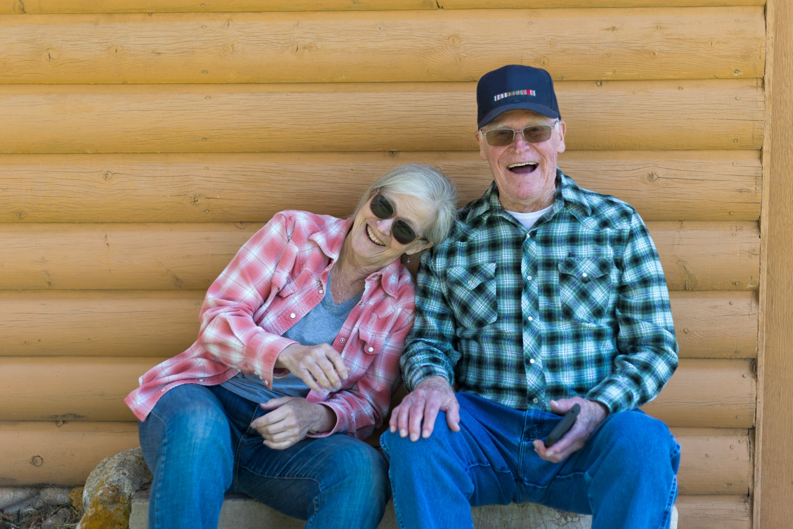 A woman and a man sitting together outdoors, smiling and enjoying each other's company against a wooden log wall.