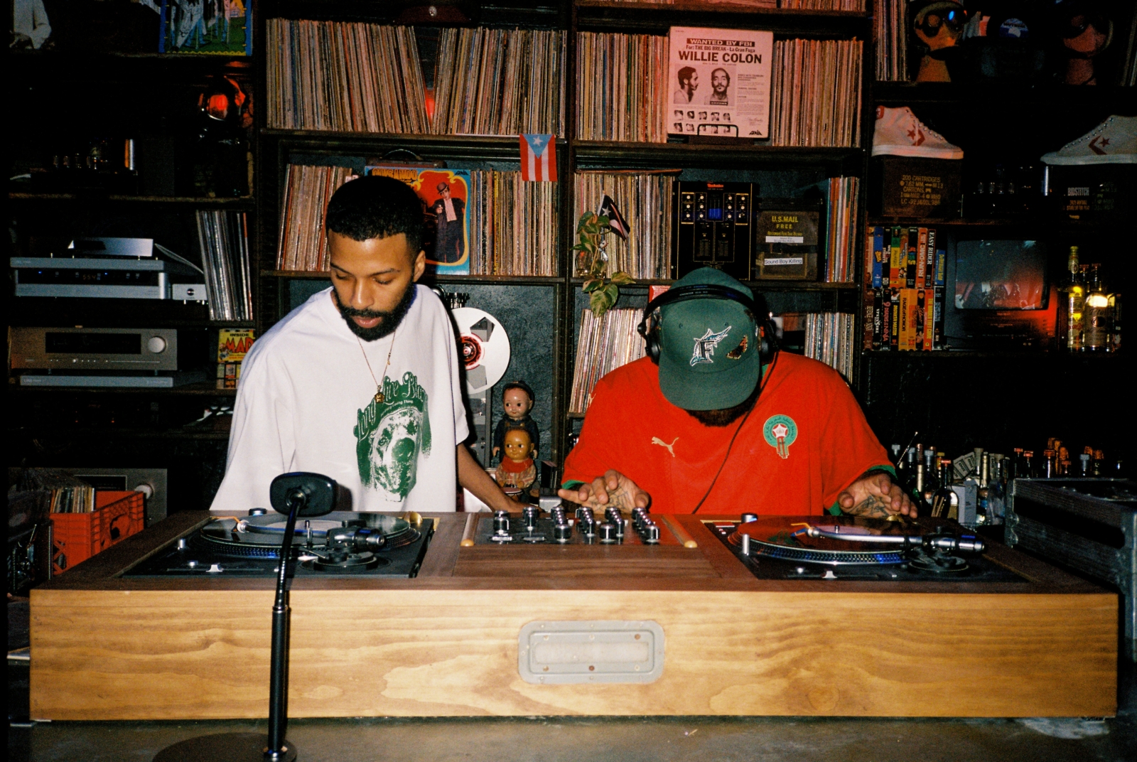 DJ duo performing with turntables in a cozy, vinyl-filled room, one wearing a white hoodie and the other in a red shirt with a cap.